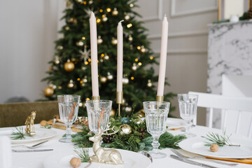 Beautiful glass glasses, candles in the decor of the festive table. Christmas table setting on the background of a Christmas tree