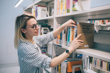Blonde woman in spectacles choosing interesting best seller from shelves in store, Caucasian...
