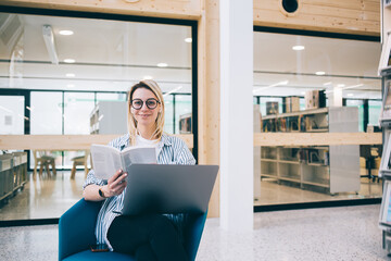 Positive woman with laptop and book in library