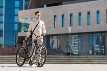 Young smiling stylish businessman pushing bicycle while going to work