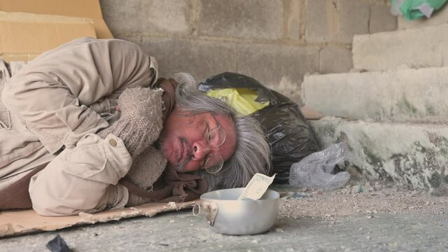 Old senior homeless man with gray beard with wear sweater and blanket sleeping the walkway street. Hand of kind woman giving banknote and money to poor man beggar. Happy elderly from money to donate.