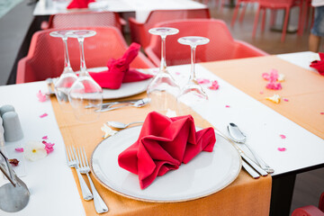 Festive table setting in the elite restaurant is ready for newlyweds. Plates, knives, forks, glasses, rose petals, red napkins, yellow tablecloth on a white table. Summer terrace. Weekend. Celebration
