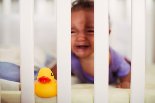 Baby crying behind the bars of her crib with toy rubber duck