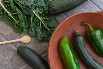 Ceramic saucepan with peppers and pickles inside, next to a wooden spoon, spinach leaf and pickles.
