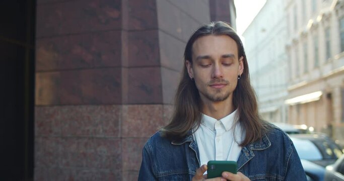 Close up view of young man using mobilephone while walking outdoors. Handsome guy with long hair scrolling and looking at phone screen at city street. Concept of real life.