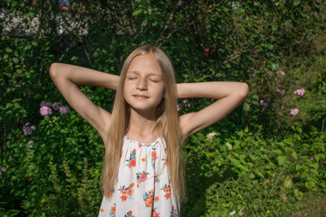 Naklejka na ściany i meble Girl with golden hair is sunbathing under the warm summer sun against a background of bright green foliage. The concept of happy childhood, sunny summer, autumn, pleasant memories and emotions