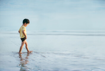 A boy walking on the shallow water at the beach as the waves calm in the morning. The surrounding atmosphere is filled with the same tone of blue in both the sky and the sea.