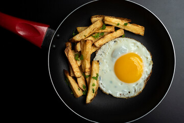 fried egg in black pan with french fries and parsley, on black background and slotted spoon and raw egg