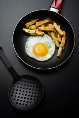 fried egg in black pan with french fries and parsley, on black background and slotted spoon and raw egg