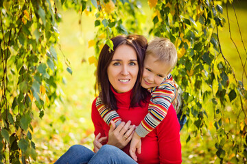 Happy family mother and son boy with sweet emotions hugging in the fall in nature, sitting on the grass near a tree.