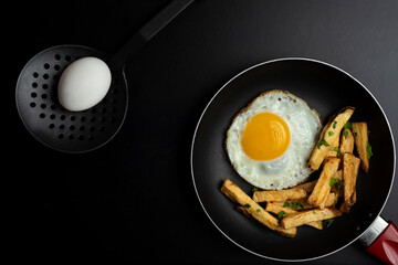 fried egg in black pan with french fries and parsley, on black background and slotted spoon and raw egg