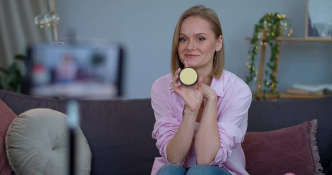 Smiling girl talking and showing to camera cosmetic product while sitting on sofa at home.Cheerful vloger recording on smartphone make up review. Concept of beauty and vlogging.