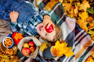 Fototapeta na wymiar Selective focus. A boy lies on a blanket with an Apple in his hands in an autumn Park. There are a lot of yellow maple leaves around. Picnic in nature in autumn.