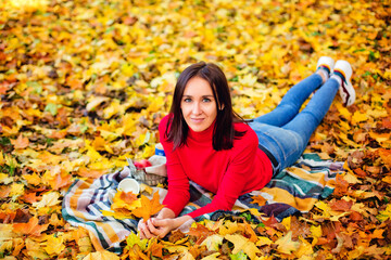 Fototapeta na wymiar A dark-haired woman in a red sweater is lying on a blanket in a Park among yellow maple leaves. Autumn picnic in nature.