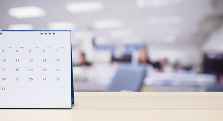 White paper desk calendar on wood table top with blurred office interior background