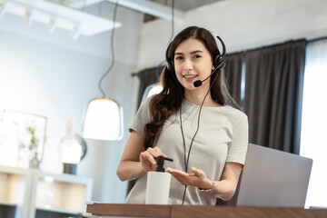 Female call operator using headphone and laptop talking with customer online while cleaning hand with alcohol gel at home. working remotely during Coronavirus or Covid19. new normal lifestyle concept 