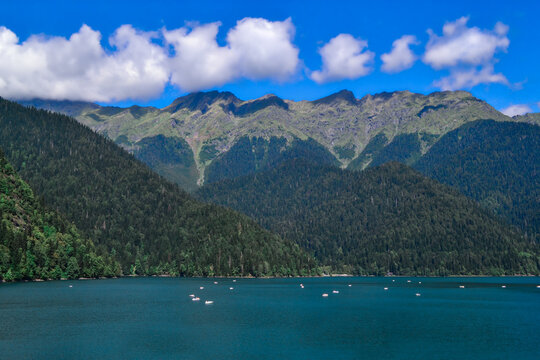 Beautiful blue clear emerald emerald Lake Ritsa glowing in the sun with ships and yaght near the middle of green trees high mountains, blue sky