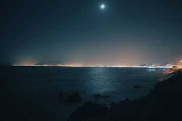 Foto del mar nocturno con reflejo de la luna y luces del puerto