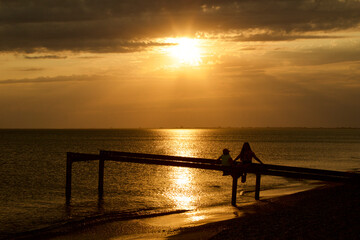 Fototapeta premium Silhouettes of two childrens figures sitting on an old pier above the sea in the rays of the setting sun. Children's friendship of two girls. The contours of children's figures against the sunset