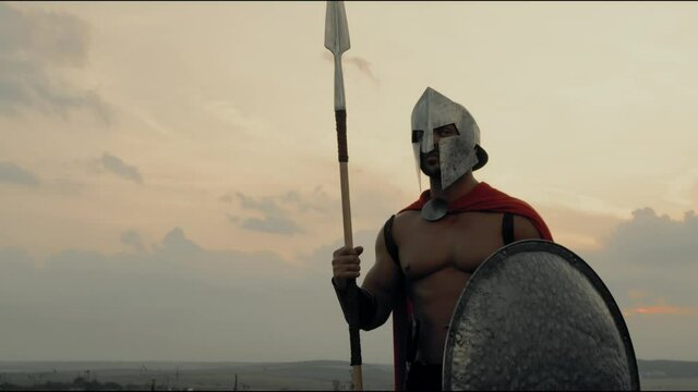 Shirtless spartan posing with spear in field.