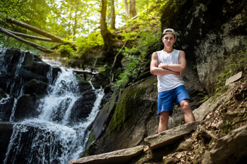 Obraz premium Handsome teenage boy is gaining strength and resting near a waterfall in the mountains, the guy likes to travel through the woods to places of interest