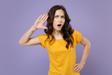 Fototapeta na wymiar Shocked curious young brunette woman 20s wearing basic yellow t-shirt posing try to hear you overhear listening intently looking aside isolated on pastel violet colour background, studio portrait.