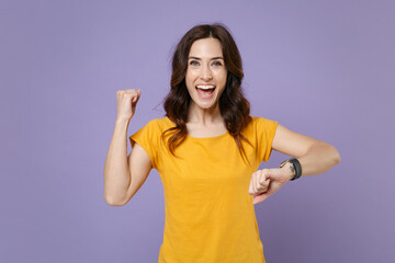 Obraz na płótnie Canvas Happy joyful young brunette woman 20s wearing basic yellow t-shirt smart watch on hand posing standing doing winner gesture looking camera isolated on pastel violet colour background, studio portrait.