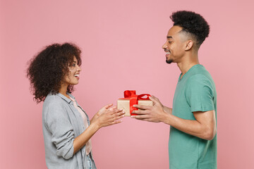 Side view smiling young african american couple friends guy girl in gray green clothes celebrating hold red present box with gift ribbon bow isolated on pastel pink color background studio portrait.