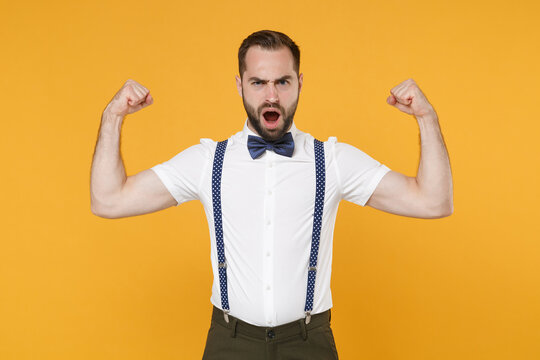 Shocked strong young bearded man 20s wearing white shirt bow-tie suspender posing showing biceps muscles keeping mouth open looking camera isolated on bright yellow color background, studio portrait.