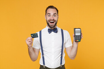 Excited young bearded man 20s wearing white shirt bow-tie suspender hold wireless modern bank payment terminal to process acquire credit card payments isolated on yellow background studio portrait.