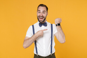 Excited funny young bearded man 20s wearing white shirt bow-tie suspender posing standing pointing index finger on smart watch on hand isolated on bright yellow color wall background studio portrait.