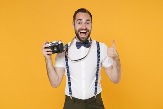 Excited young bearded man 20s wearing white shirt bow-tie suspender posing taking picture with retro vintage photo camera showing thumb up isolated on bright yellow color background studio portrait.