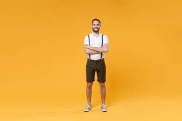 Fototapeta na wymiar Full length portrait of smiling young bearded man 20s wearing white shirt suspender shorts posing standing holding hands crossed looking camera isolated on bright yellow color wall background studio.