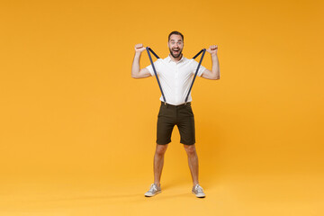 Fototapeta na wymiar Full length portrait of excited shocked young bearded man 20s wearing white shirt shorts posing stretching suspender keeping mouth open looking camera isolated on bright yellow wall background studio.
