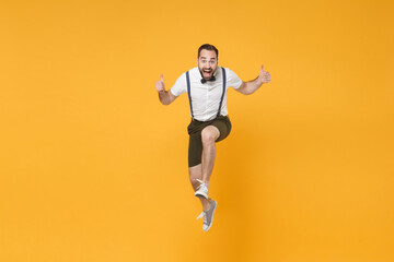 Fototapeta na wymiar Full length portrait of excited young bearded man 20s wearing white shirt suspender shorts posing jumping showing thumbs up looking camera isolated on bright yellow color wall background studio.