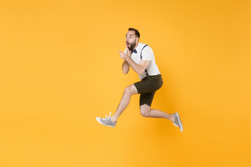Fototapeta na wymiar Full length side view portrait of shocked young bearded man 20s in white shirt suspender shorts posing jumping pointing index fingers aside on mock up copy space isolated on yellow background studio.