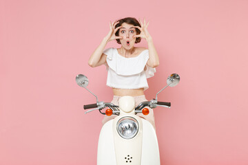 Shocked amazed young woman 20s in white summer clothes posing stretching eyelids keeping mouth open looking camera sitting driving moped isolated on pastel pink colour background studio portrait.