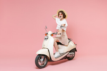 Fototapeta na wymiar Full length portrait of excited young brunette woman wearing white summer clothes hat glasses keeping mouth open looking aside sitting driving moped isolated on pastel pink colour background studio.