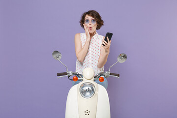Shocked young brunette woman 20s in white dotted shirt glasses using mobile cell phone typing sms message put hand on cheek driving moped isolated on pastel violet colour background studio portrait.