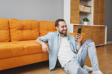 Smiling cheerful young bearded man wearing casual white t-shirt blue shirt using mobile cell phone typing sms message sitting at floor near couch resting relaxing spending time in living room at home.