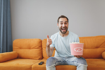 Cheerful funny amazed young bearded man wearing casual blue t-shirt watching movie film, holding bucket of popcorn showing thumb up sitting on couch resting spending time in living room at home.