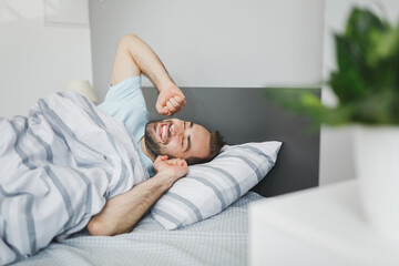 Smiling attractive young bearded man wearing basic blue t-shirt sleeping waking up stretching hands lying in bed with striped sheet pillow blanket resting relaxing spending time in bedroom at home.