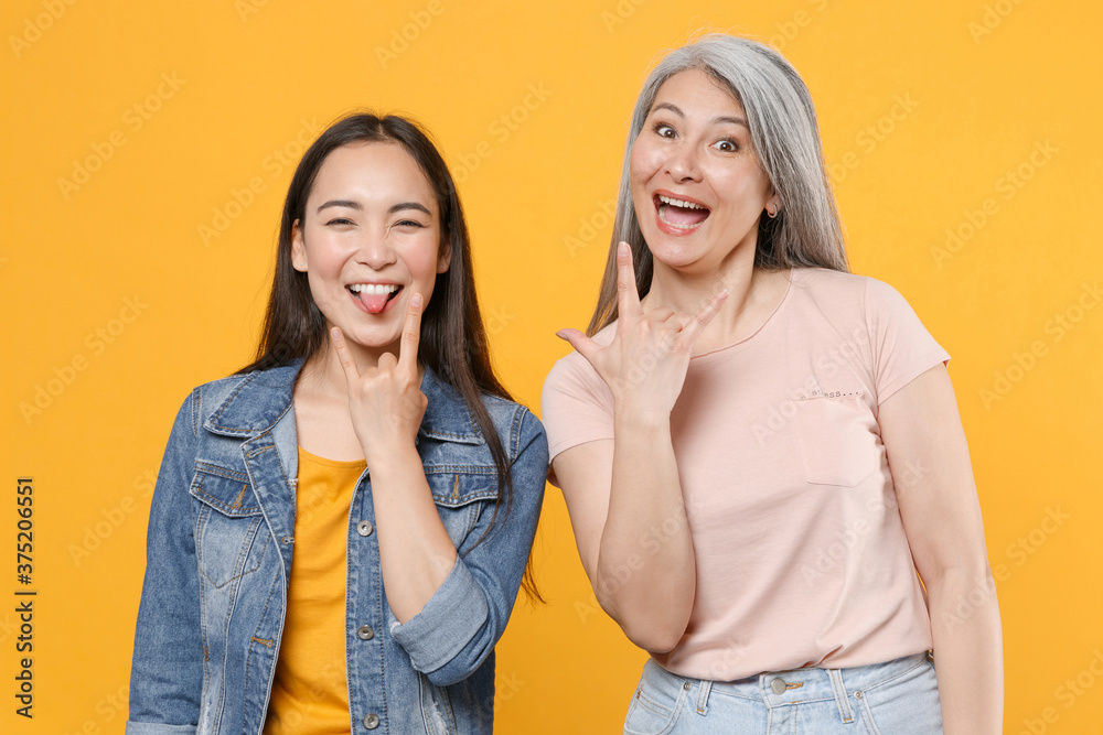Wall mural Cheerful family asian women girls gray-haired mother brunette daughter in casual clothes posing depicting heavy metal rock sign horns up gesture isolated on yellow color background studio portrait. - Wall murals