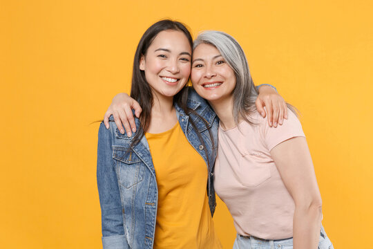 Smiling cheerful joyful family asian female women girls gray-haired mother and brunette daughter in casual clothes posing hugging looking camera isolated on yellow color background studio portrait.