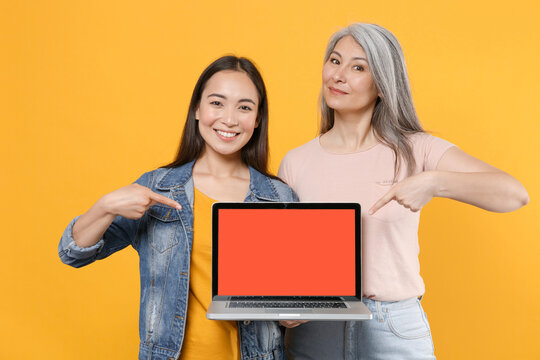 Smiling family asian women girls mother daughter in casual clothes point index finger on laptop pc computer with blank empty screen mock up copy space isolated on yellow background studio portrait.