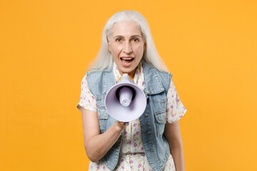 Excited cheerful elderly gray-haired female woman 60s 70s wearing casual dress denim waistcoat posing screaming in megaphone looking camera isolated on yellow color wall background studio portrait.