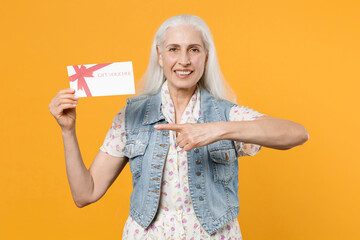 Smiling elderly gray-haired female woman wearing casual dress denim waistcoat posing pointing index finger on gift certificate looking camera isolated on yellow color wall background studio portrait.