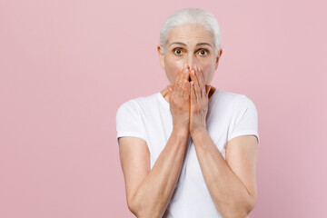 Shocked elderly gray-haired female woman 60s 70s wearing white blank design casual t-shirt posing covering mouth with hands looking camera isolated on pastel pink color background studio portrait.