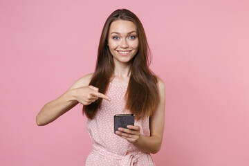 Smiling young brunette woman 20s wearing pink summer dotted dress posing point index finger on mobile cell phone typing sms message looking camera isolated on pastel pink background studio portrait.