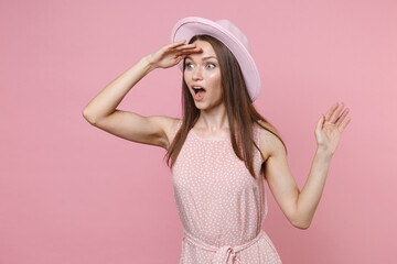Shocked young brunette woman 20s in pastel pink summer dotted dress hat posing holding hand at forehead looking far away distance spreading hands isolated on pastel pink background studio portrait.